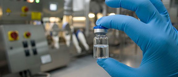 Hand in blue gloves holding a pharmaceutical vial with equipment in the background.